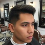 Deskripsi gambar: Potongan rambut pria low fade 2 jari dengan gradasi halus mulai dua jari di atas telinga, samping dan belakang memudar ke kulit, bagian atas textured pendek sekitar 4-5 cm, ditata messy natural. Tampak samping dan depan, pria berwajah tegas dengan kulit sawo matang, latar belakang barbershop modern.