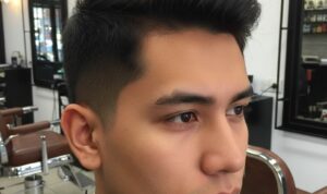 Deskripsi gambar: Potongan rambut pria low fade 2 jari dengan gradasi halus mulai dua jari di atas telinga, samping dan belakang memudar ke kulit, bagian atas textured pendek sekitar 4-5 cm, ditata messy natural. Tampak samping dan depan, pria berwajah tegas dengan kulit sawo matang, latar belakang barbershop modern.